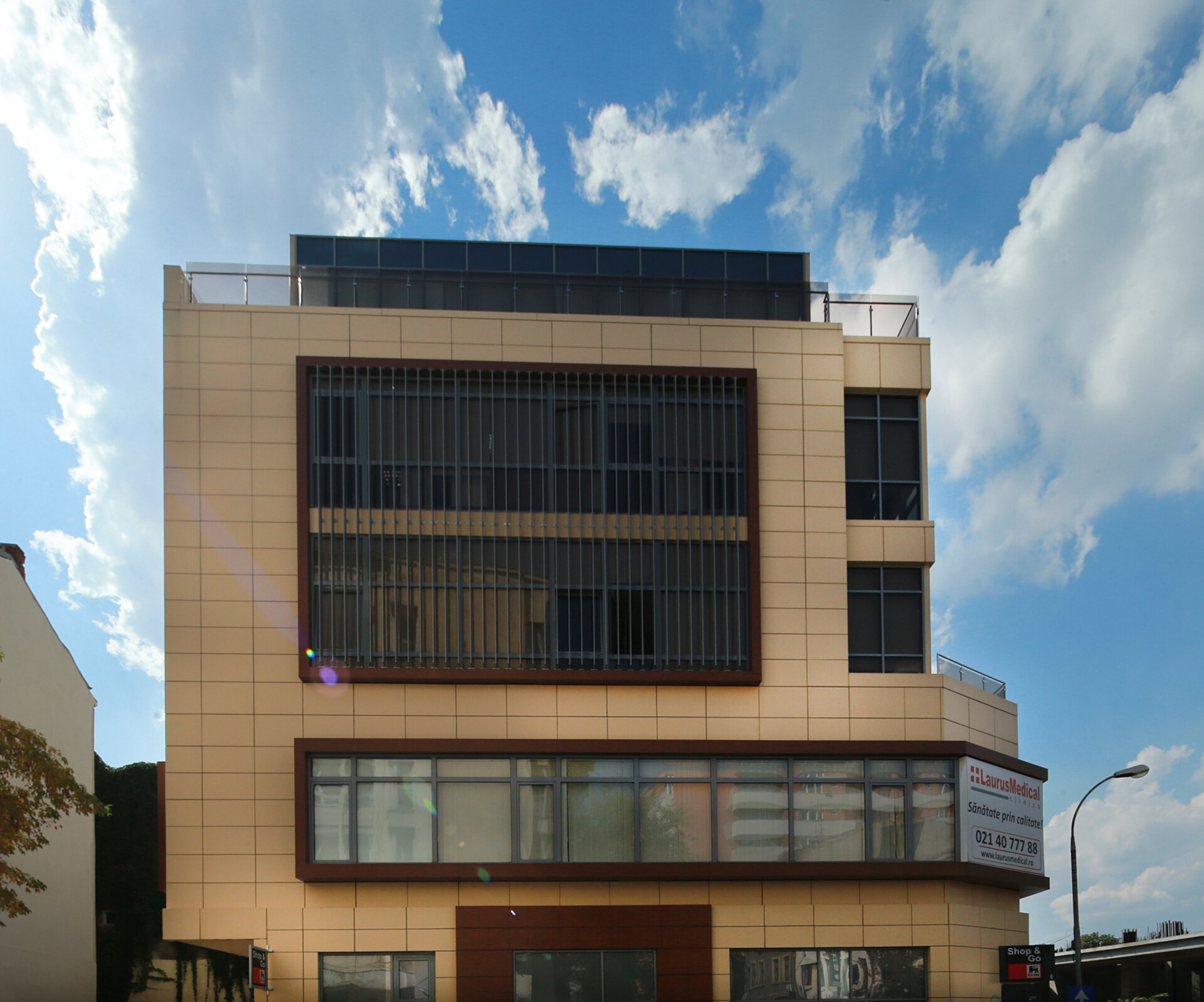 Fațadă principală clădire Mihai Eminescu 100 cu placaj ceramic în tonuri calde, parasolare verticale pe etajul superior, finisaj profil-lemn pe paneluri orizontale și siglă „Laurus Medical Clinic” pe parter — clădire S+P+4E, proiect PPAE, faza inițială de policlinică